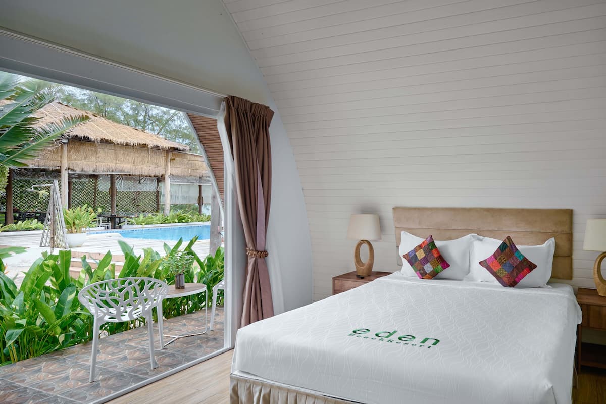 Una habitación de hotel con cama, mesita de noche, ventana con cortinas, vista a una piscina y un patio con una silla y plantas.