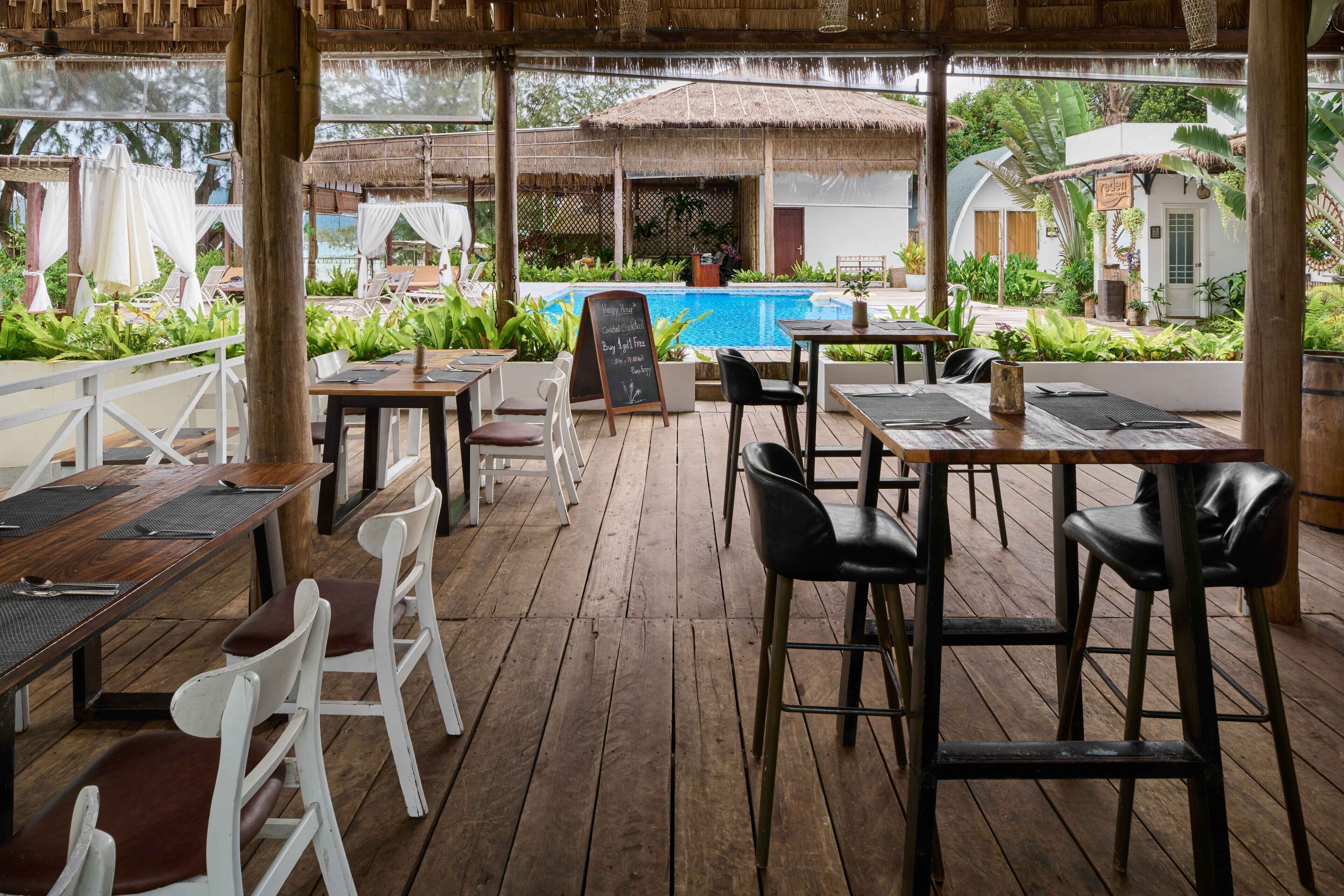 An outdoor dining area with wooden tables and chairs, a pool in the background, and lush greenery.