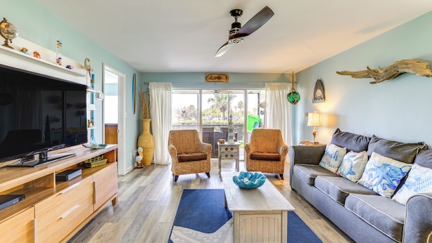 A living room with a TV, wooden entertainment unit, two wicker chairs, a grey sofa, and a ceiling fan. - Beachfront Siesta Key Condo w/ Enclosed Patio (Siesta Key)