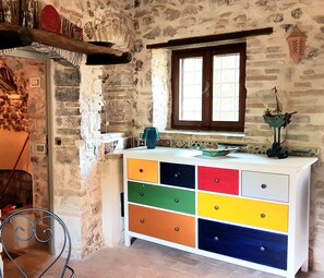 Una habitación con pared de piedra, ventana de madera, cómoda de colores y una pequeña mesa con una silla.