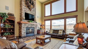 A spacious living room with a stone fireplace, leather sofas, and large windows offering a mountain view. - Riverview Retreat: Incredible Views, Heated Pool, Spa, Theater Room & More! (East Wenatchee)