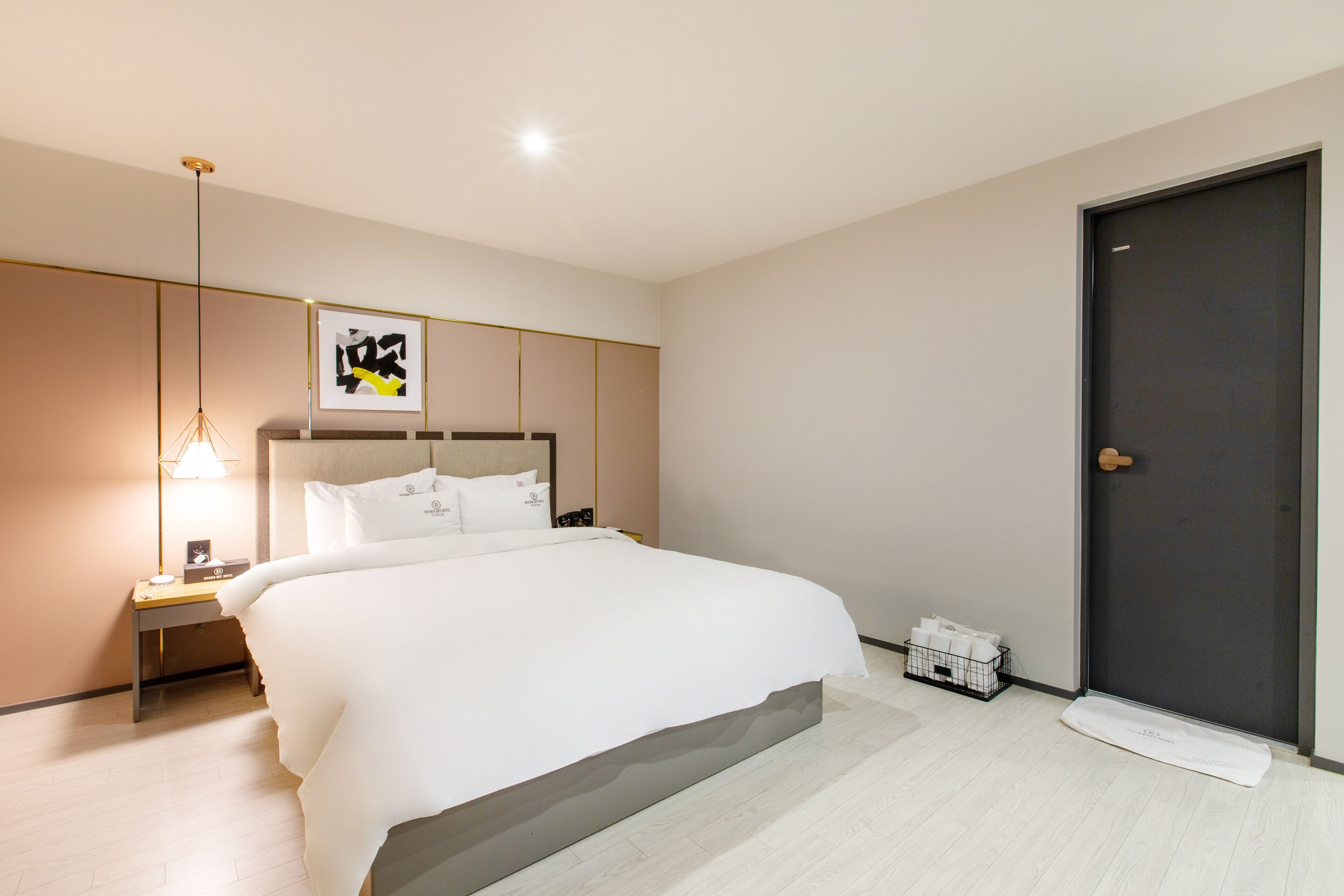 A modern hotel room with a large bed, a bedside table with a lamp, a framed artwork on the wall, and a door to the right.