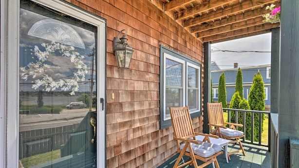 A wooden porch with two wooden chairs and a table, a window with a decorative wreath, and a view of a road and houses in the background.