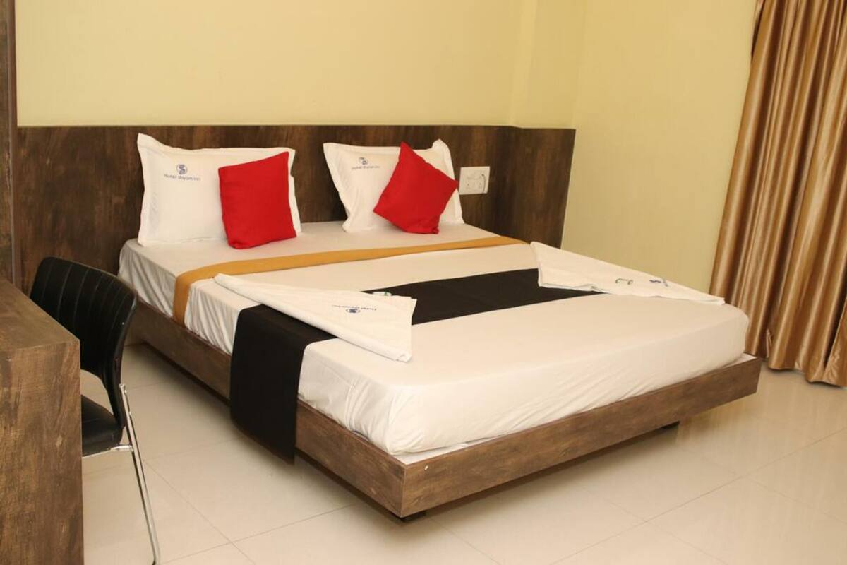 A hotel room with a wooden bed frame, white bedding, red pillows, and a black throw blanket.