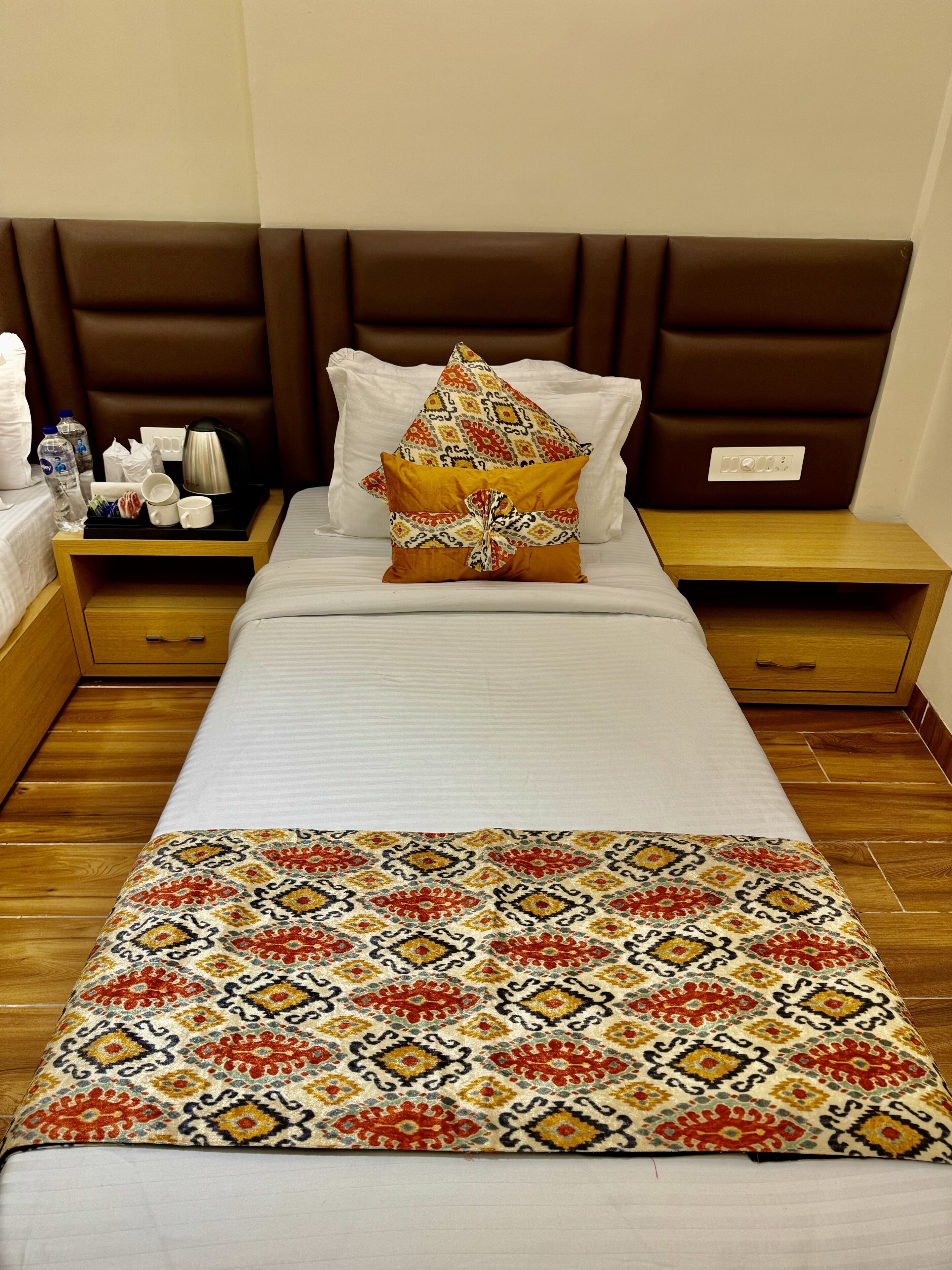 A neatly made bed with a patterned bedspread, a brown headboard, and a bedside table with a lamp and toiletries.