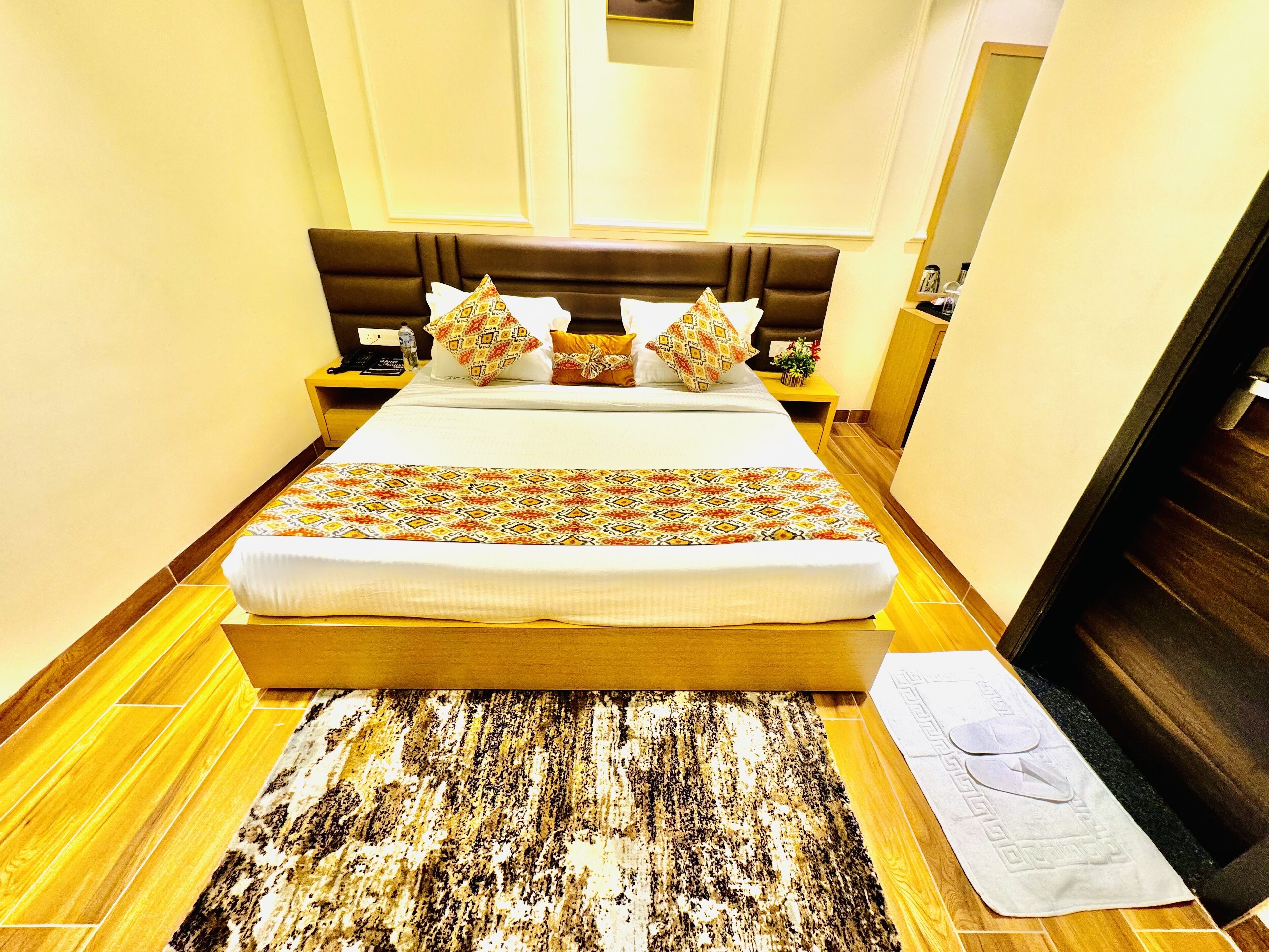 A neatly made bed with a patterned bedspread, a brown headboard, and multiple pillows. There is a wooden nightstand with a lamp and a bottle of water. The room has a wooden floor and a small rug.