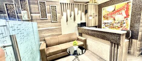 A reception area with a wooden reception desk, a brown sofa, a glass coffee table with flowers, and patterned wall decorations.