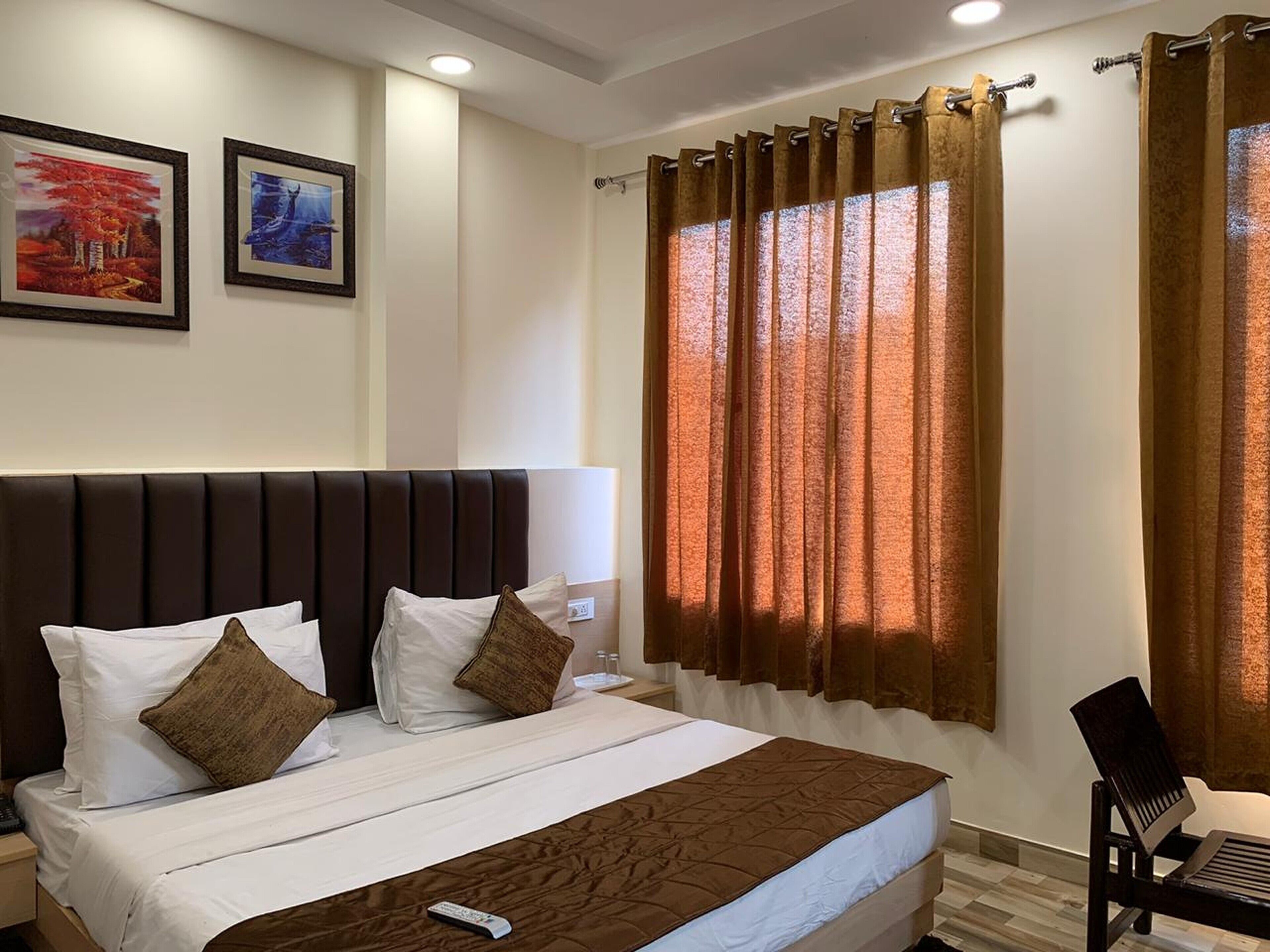 A hotel room with a large bed, a headboard, two pillows, a remote control, a chair, and curtains.