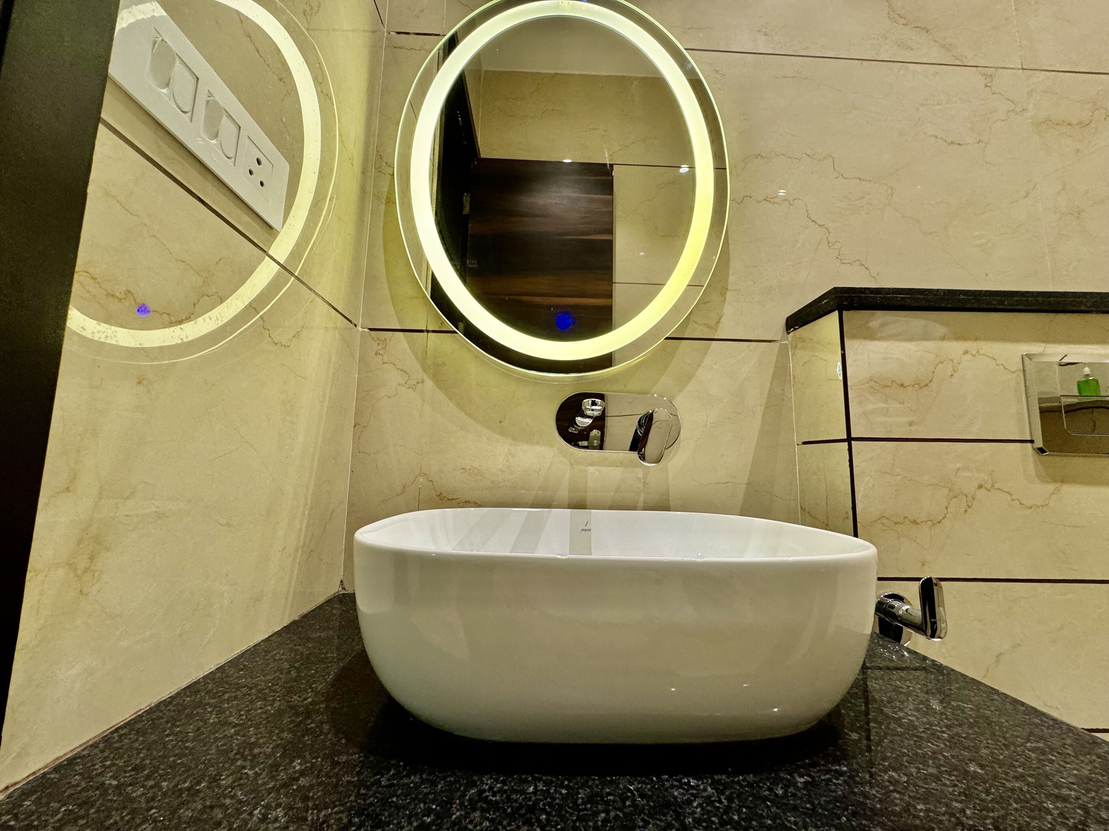 Une salle de bain moderne avec un grand lavabo ovale, un miroir rond avec un éclairage intégré et une petite étagère contenant des articles de toilette.