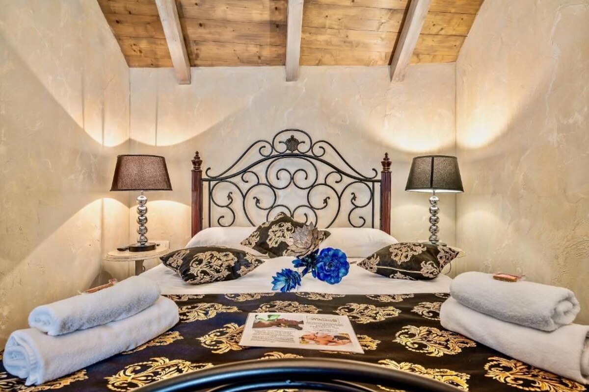 A bedroom with a bed, two bedside lamps, a decorative pillow, a blue flower, and rolled towels.
