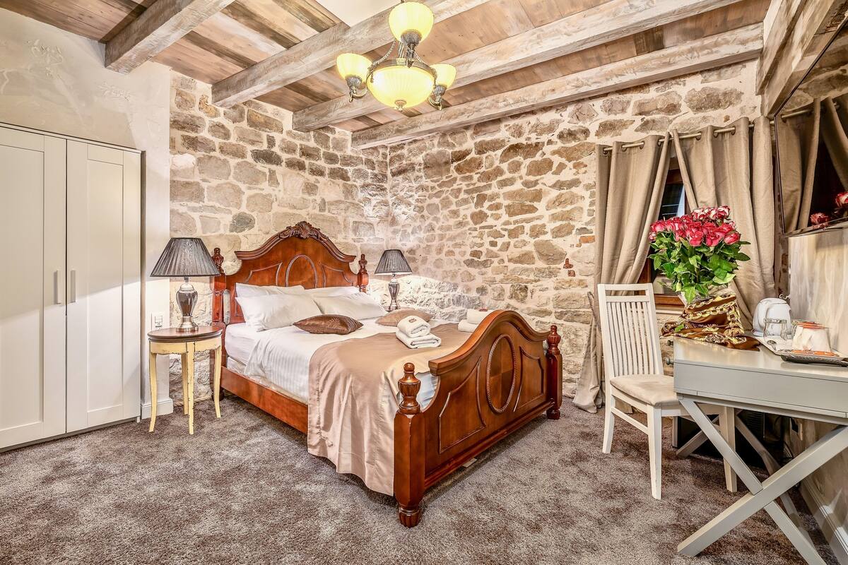 A bedroom with a wooden bed, a desk, and a chair. There is a stone wall and a wooden ceiling.