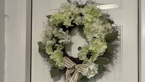 Una puerta blanca con un adorno de flores y hojas verdes, atado con un lazo a rayas, y un picaporte.