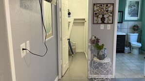 Un pasillo con un televisor montado, una puerta blanca que da a una habitación, una mesita con una planta y un baño visible a través de una puerta entreabierta.