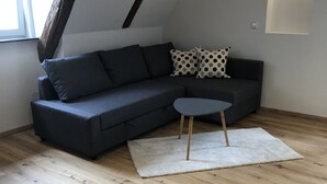 Un salon moderne avec un canapé d’angle gris foncé, une petite table ronde et un tapis blanc. La pièce est dotée d’un plancher en bois et d’un plafond en pente avec des poutres apparentes.