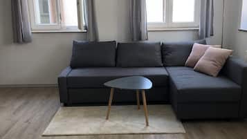 Ein modernes Wohnzimmer mit einem dunkelgrauen Ecksofa, einem kleinen runden Tisch und einem weißen Teppich. Zwei Fenster mit grauen Vorhängen sind zu sehen, außerdem eine hohe Stehlampe.