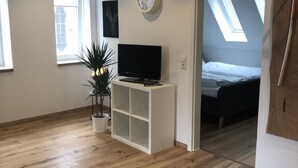Une chambre moderne avec un plancher en bois, un meuble TV blanc, une plante en pot, une horloge murale et un lit dans la pièce adjacente.