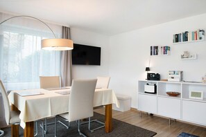 Moderni hotellihuone, jossa on ruokapöytä, tuoleja, televisio, kirjahyllyjä ja yöpöytä.