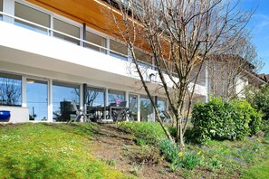 Un edificio moderno con amplios ventanales, un piso superior de madera y un jardín ajardinado con diversas plantas y flores.