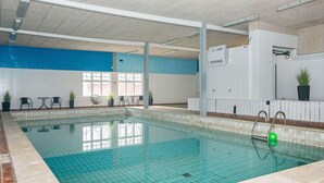 An indoor swimming pool with clear blue tiles, a brick border, and a white wall with a blue section. There are chairs and tables outside the pool area, and a potted plant on the wall. - 6 Person Holiday Home in Hojer (Højer)