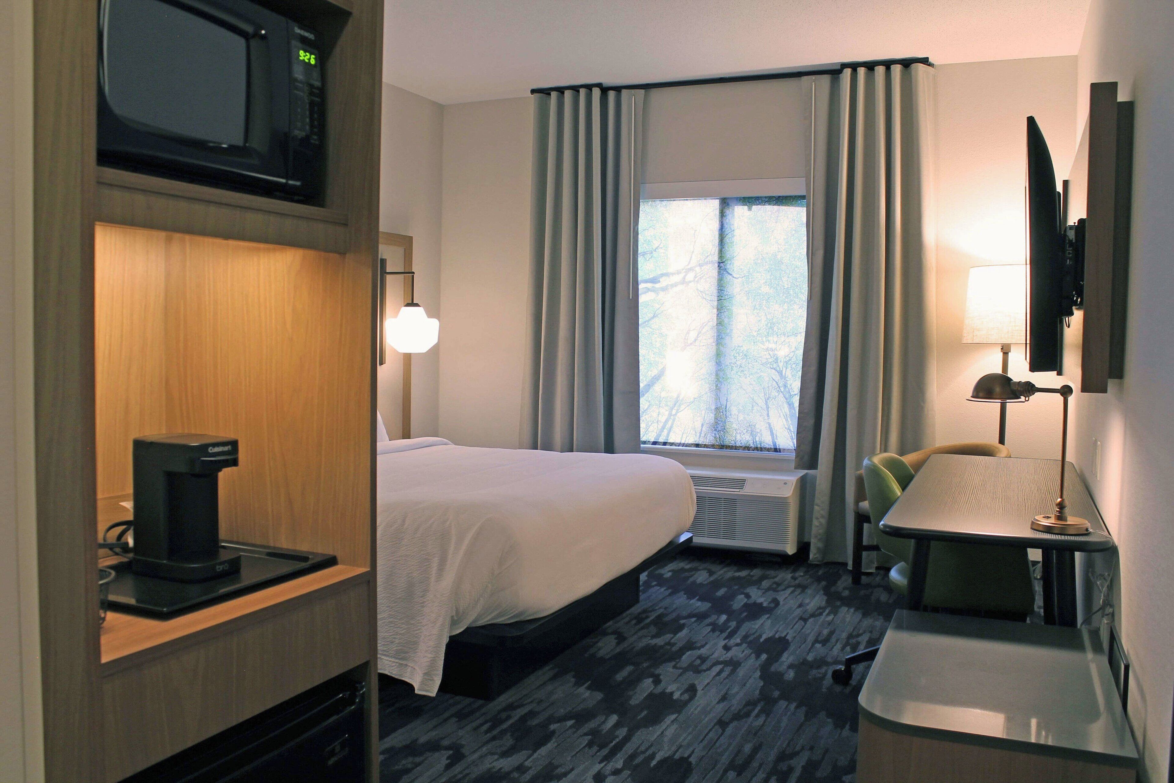 A hotel room with a bed, a desk, a coffee maker, a TV, and a window with curtains.