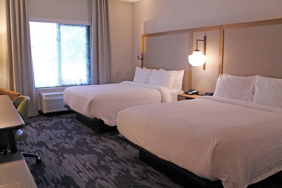Una habitación de hotel con dos camas, un escritorio, una silla, una ventana con cortinas y una lámpara en la pared.