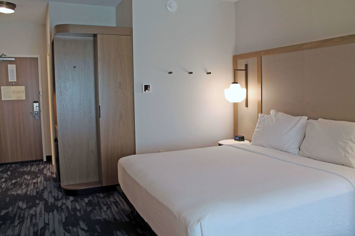 Una habitación de hotel con una cama grande, un armario de madera, una mesita de noche con lámpara y una lámpara de pared.