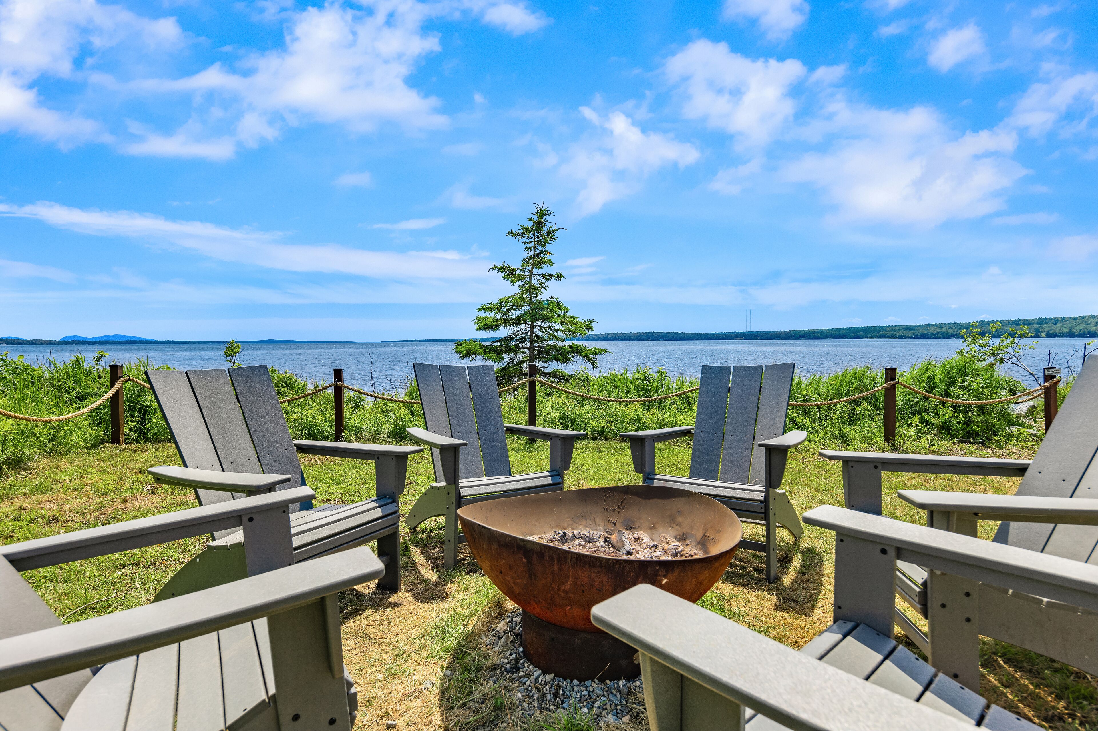 A row of Adirondack chairs facing a large metal fire pit with a view of a lake and a tree in the background.