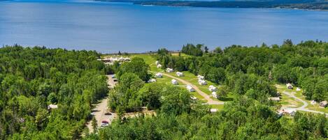 An aerial view of a campsite with tents and cabins surrounded by dense forest, with a lake in the background.