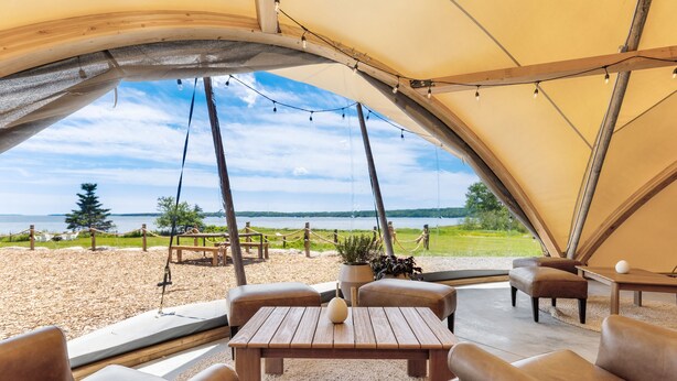 Tienda de campaña con mesa de madera y sillas de cuero, con vista a una playa y al agua.