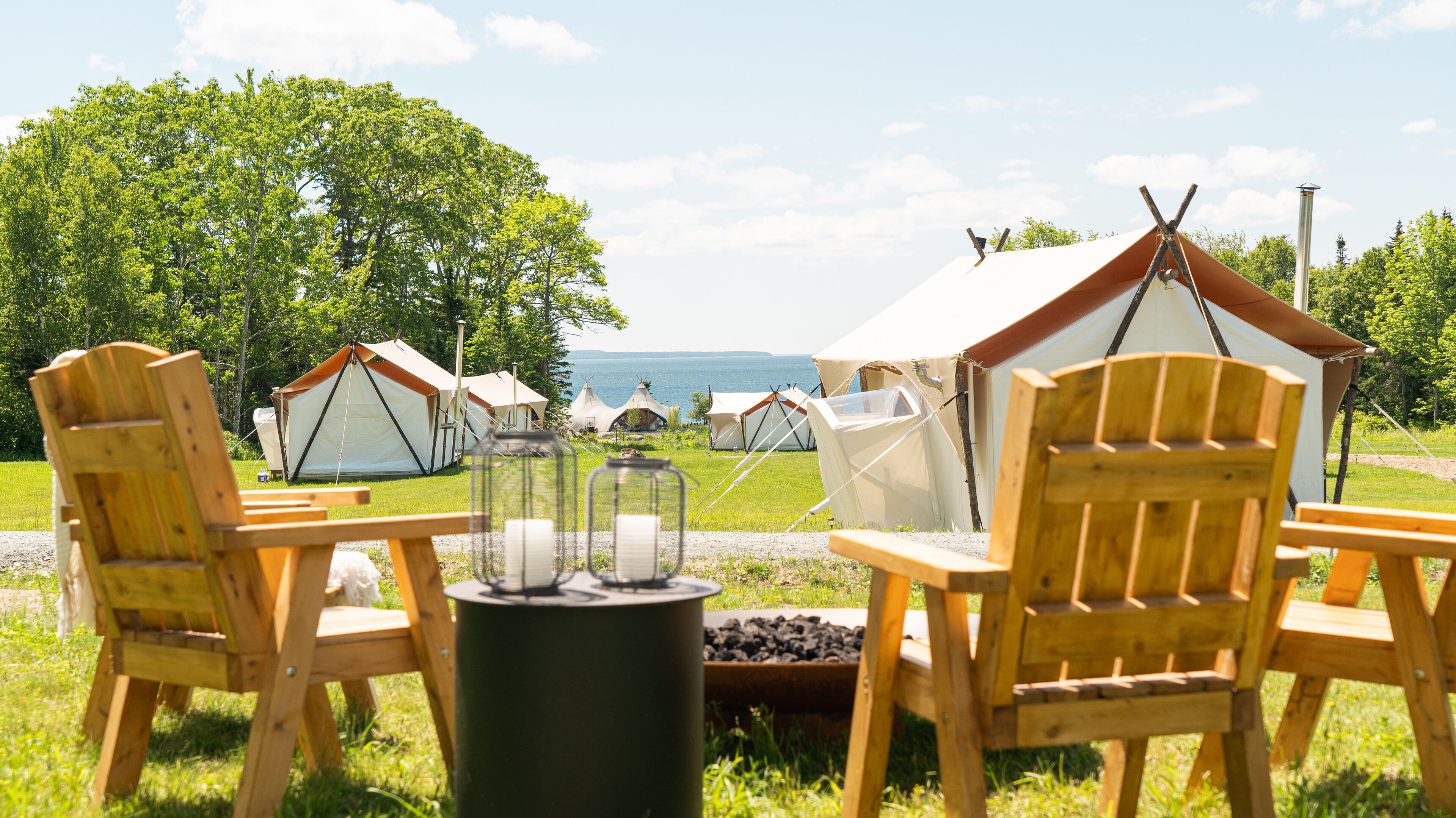 Um camping com cadeiras e mesa de madeira, tendas e vista para o mar.
