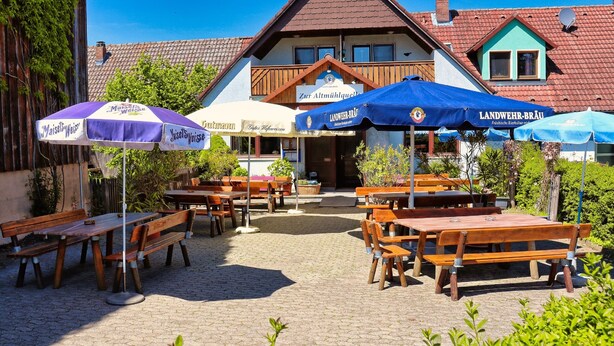 An outdoor seating area with wooden tables and benches, surrounded by potted plants and umbrellas. - Gasthof Zur Altmühlquelle (Windelsbach)