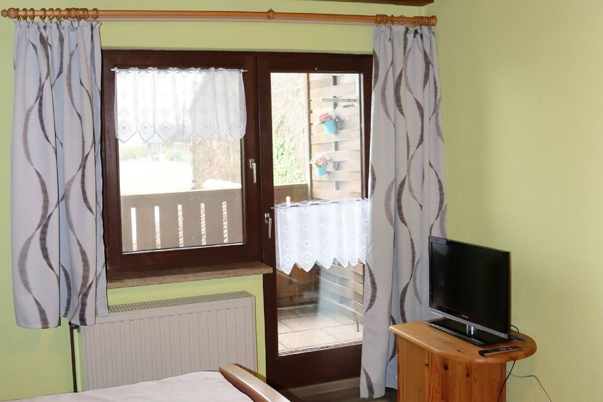 A wooden ceiling, a bed with a mattress, a television on a wooden stand, and a window with curtains.