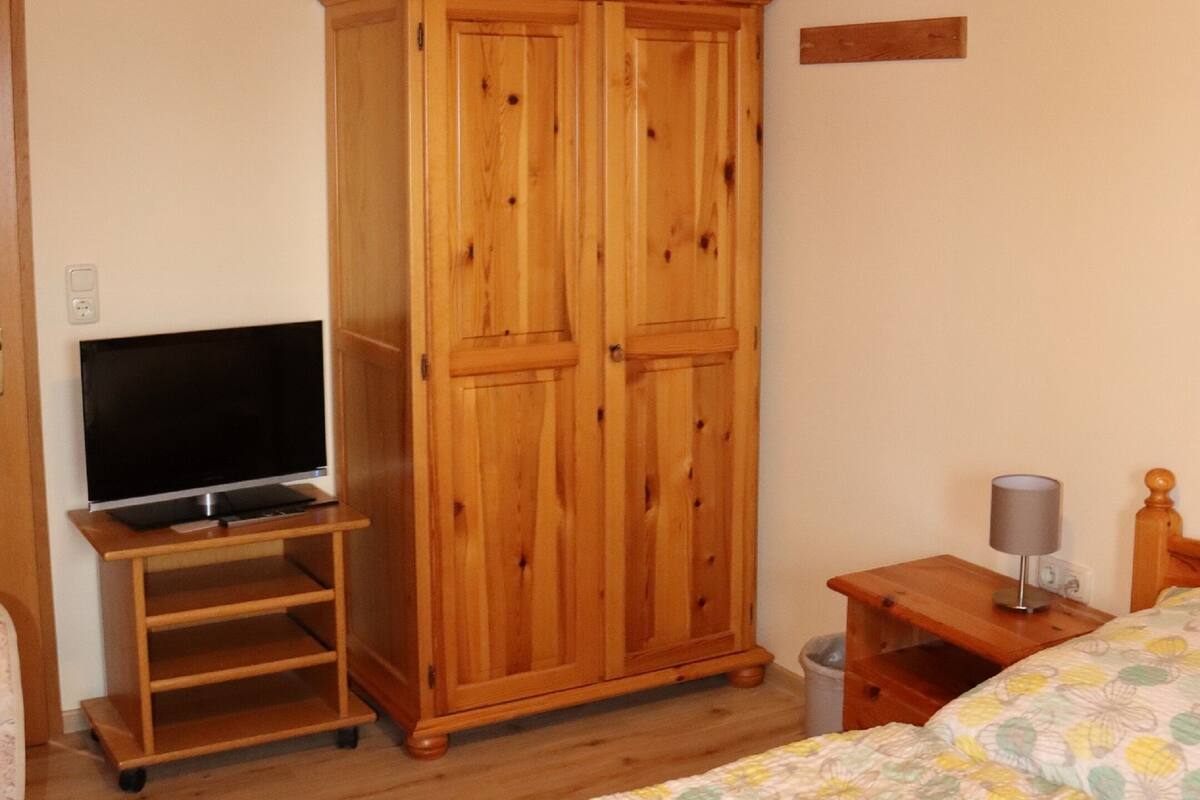 A bedroom with a wooden wardrobe, a television on a stand, a bedside table with a lamp, and a bed with a floral bedspread.