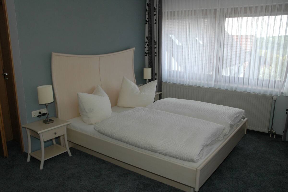 Una cama doble con ropa de cama blanca, una mesita de noche con lámpara y una ventana con cortinas translúcidas.