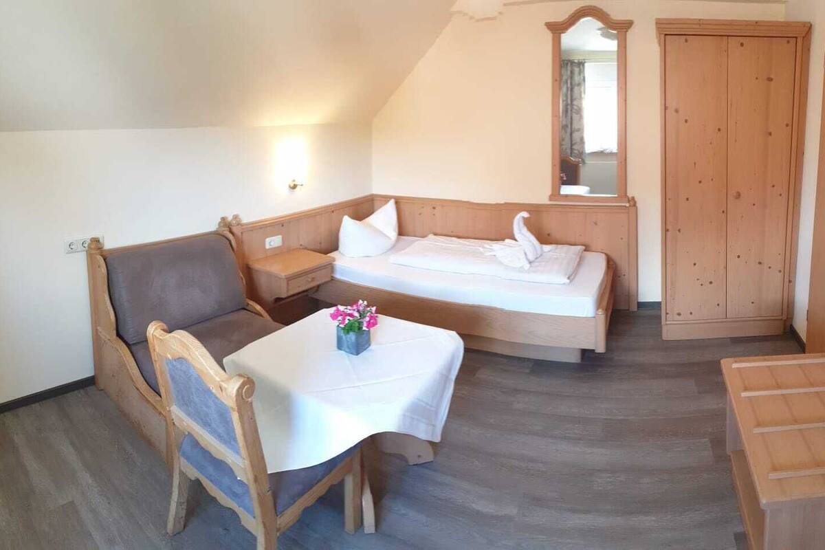 Una habitación acogedora con una cama, una mesita con flores, una silla, una mesita de noche, un armario de madera y un espejo.