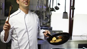 A chef holding a pan with food, in a kitchen with hanging utensils. - Hotel Zur Igelstadt (Lichtenfels)