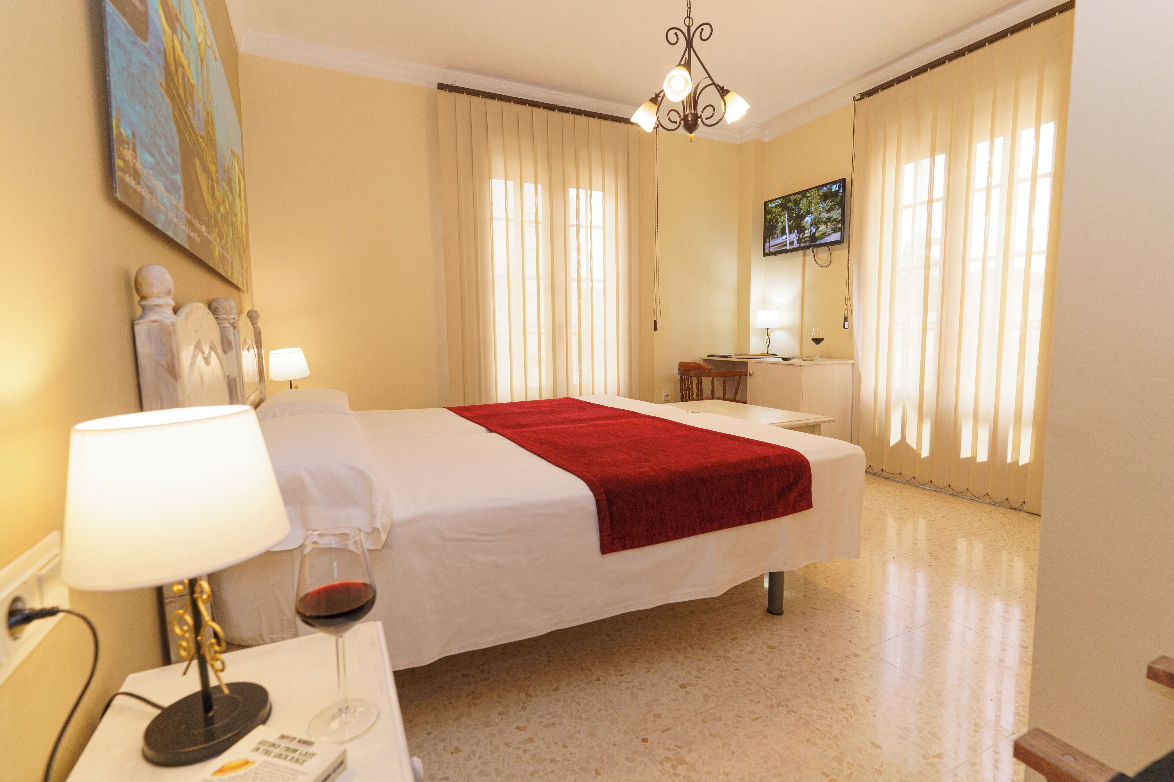 A hotel room with a bed, a bedside table with a lamp, a wine glass, and a painting on the wall.