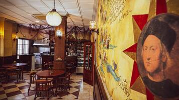 A restaurant interior with a checkered floor, wooden tables, and chairs. There is a kitchen area with a stove and a refrigerator.