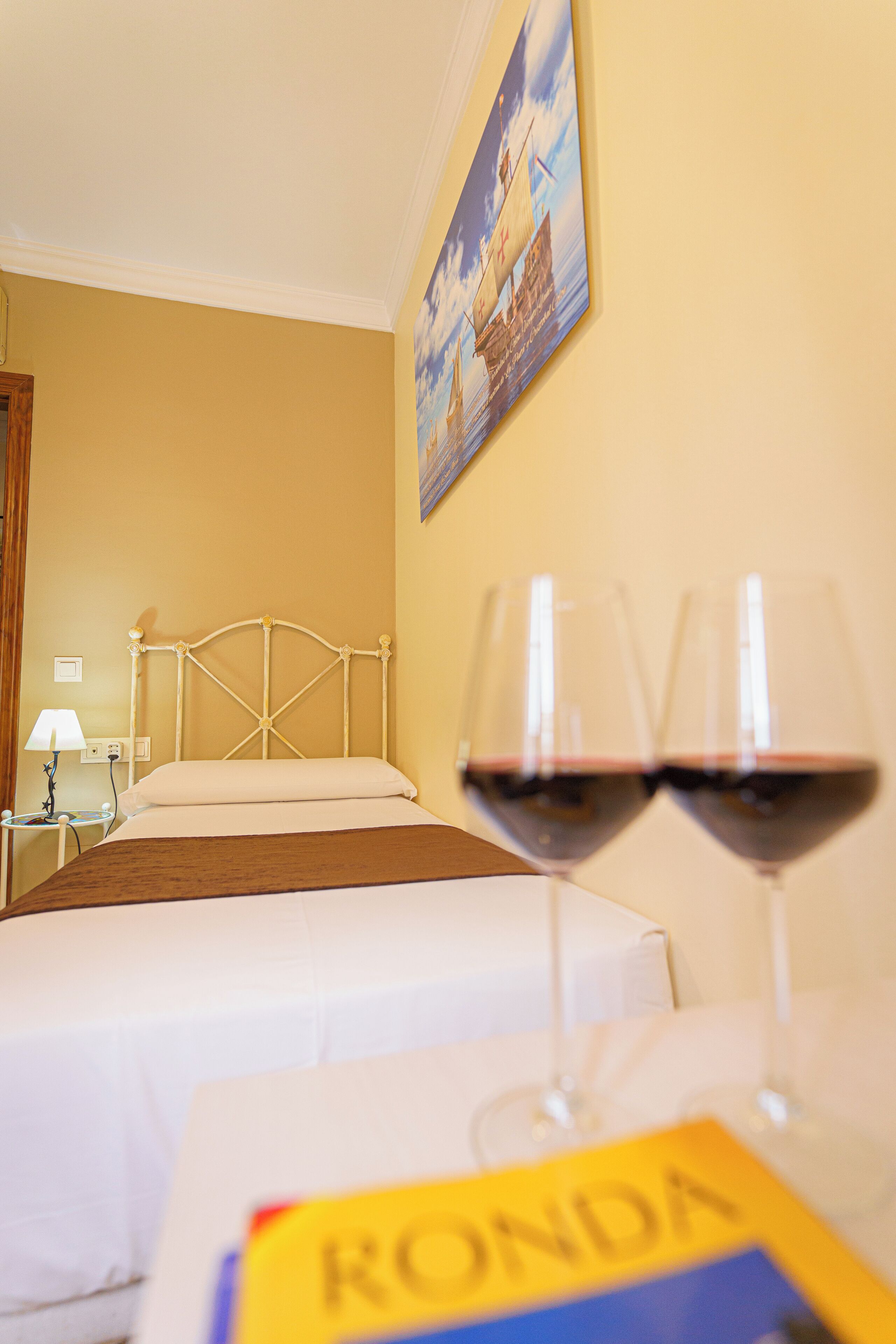 A hotel room with a bed, a nightstand, a lamp, and two wine glasses on a table.
