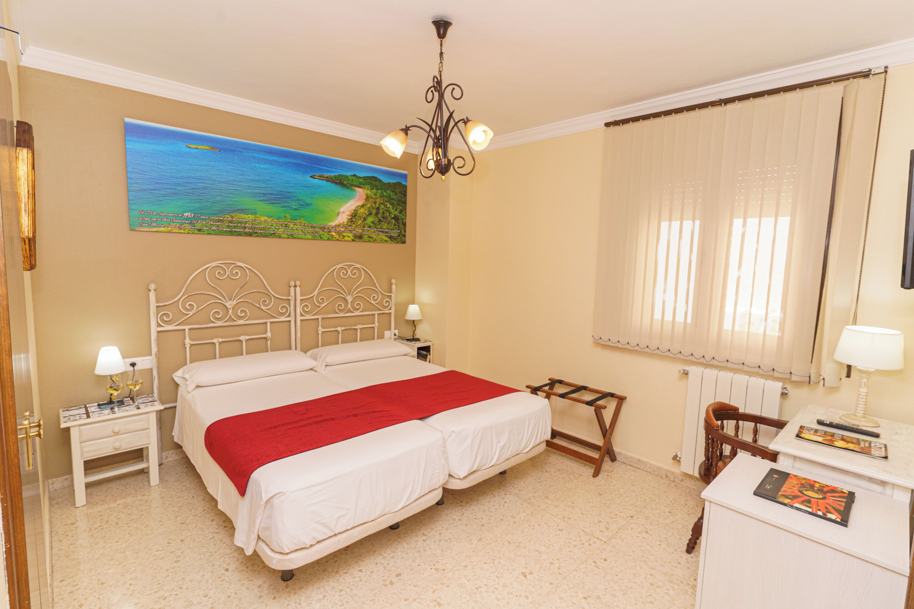 A bedroom with a large bed, a painting of a beach, a chandelier, a window with curtains, and a desk with a lamp.