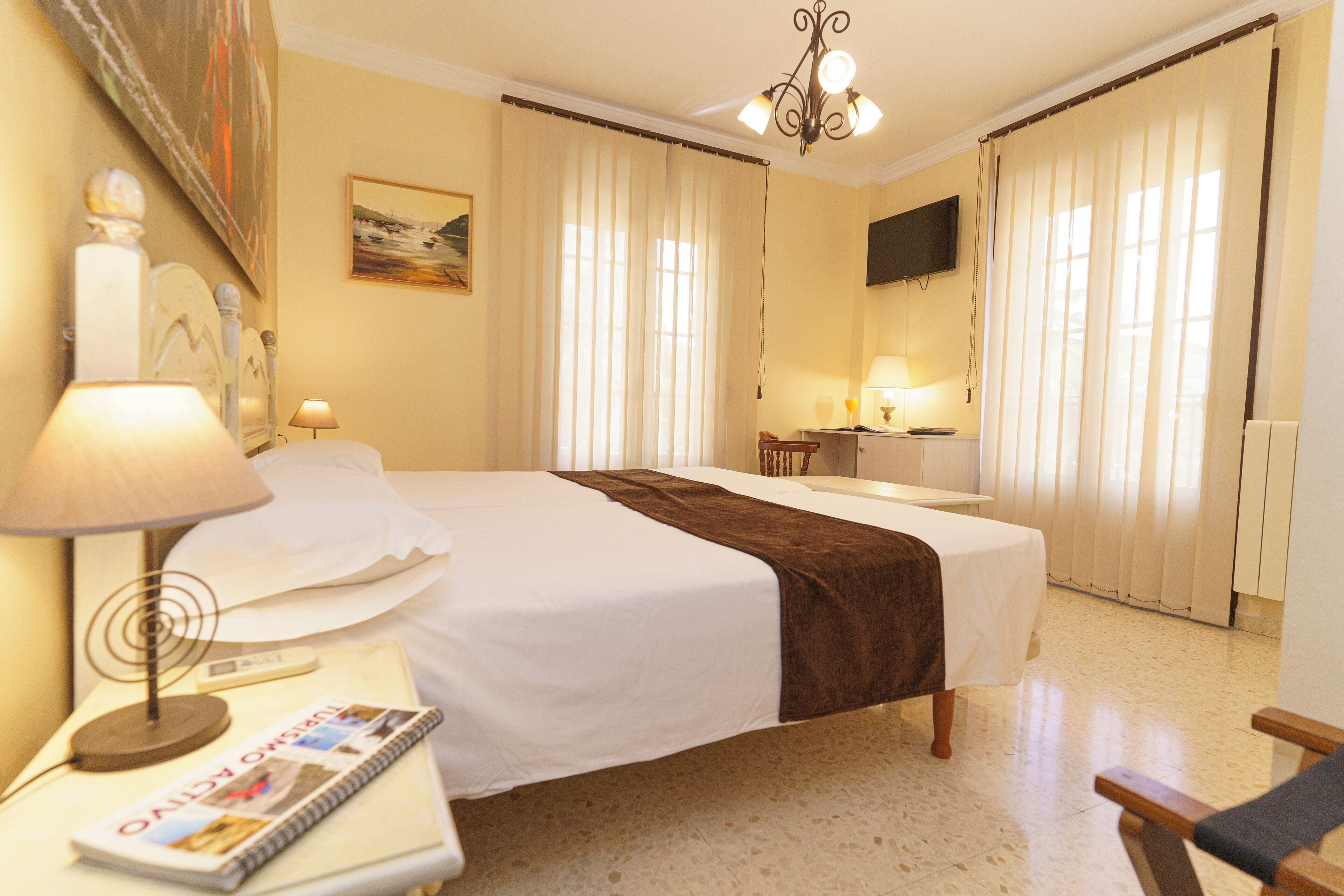 Habitación de hotel con cama, lámparas de noche, escritorio con lámpara, una silla, televisor y cortinas.