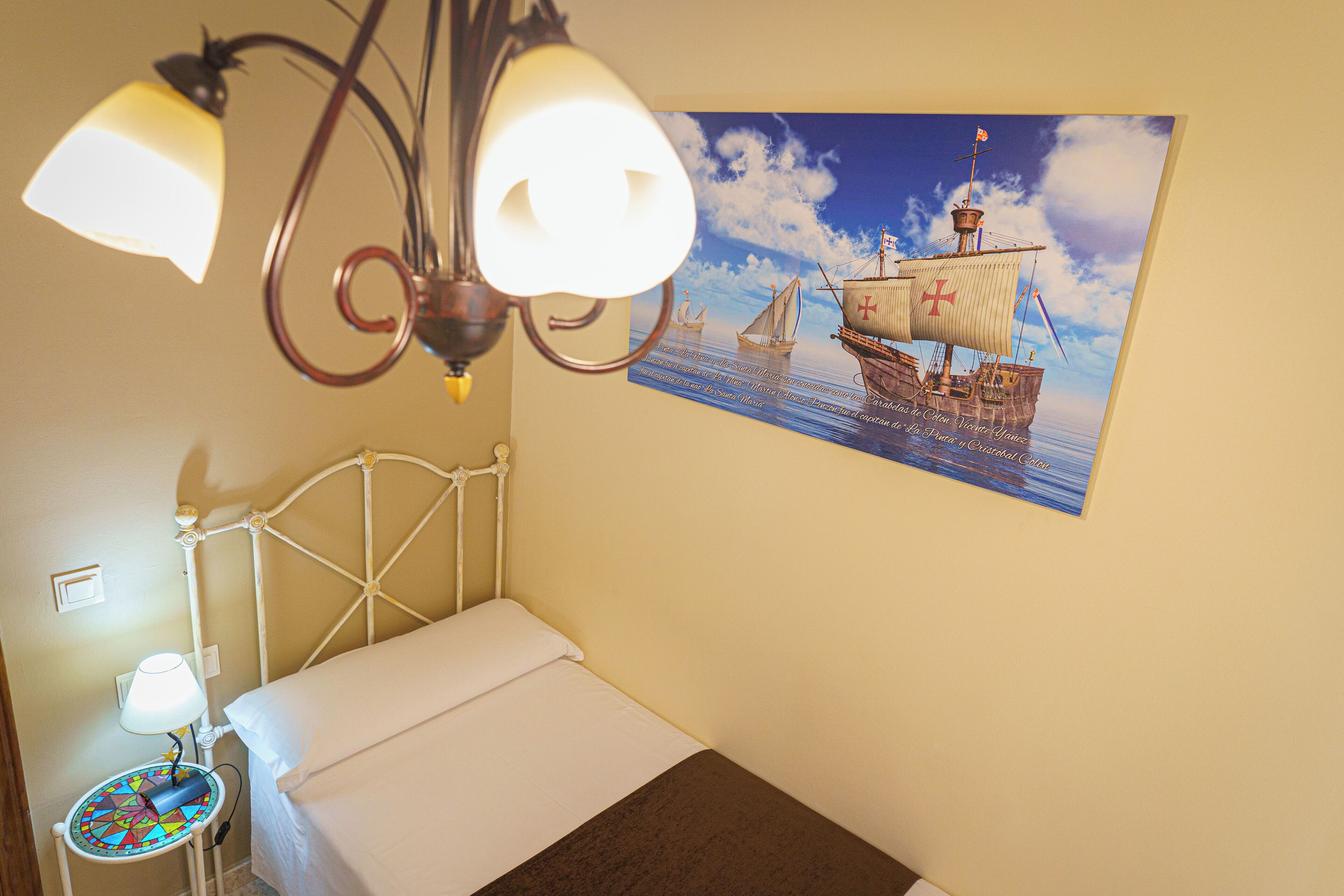 Una cama pequeña y bien hecha con cabecera blanca, una mesita de noche con una lámpara y un cuadro colgado en la pared de un barco de vela.