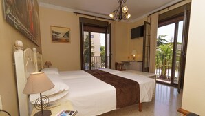 A hotel room with two beds, a desk with a lamp, a chair, and a balcony with a view of palm trees. - Hotel Colón (Ronda)