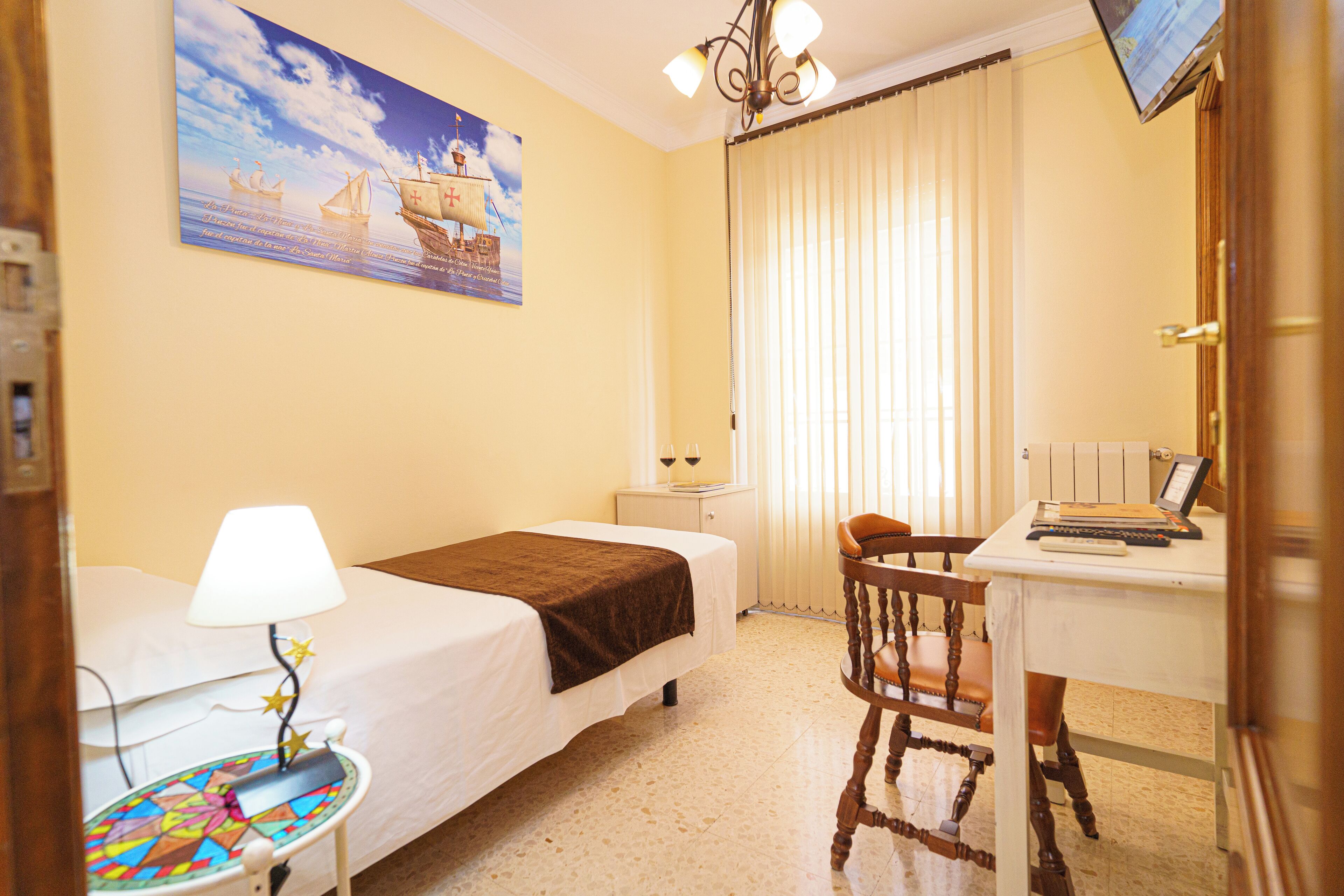Una habitación de hotel con cama, escritorio con laptop, silla, mesita con lámpara y un cuadro de un barco en la pared.