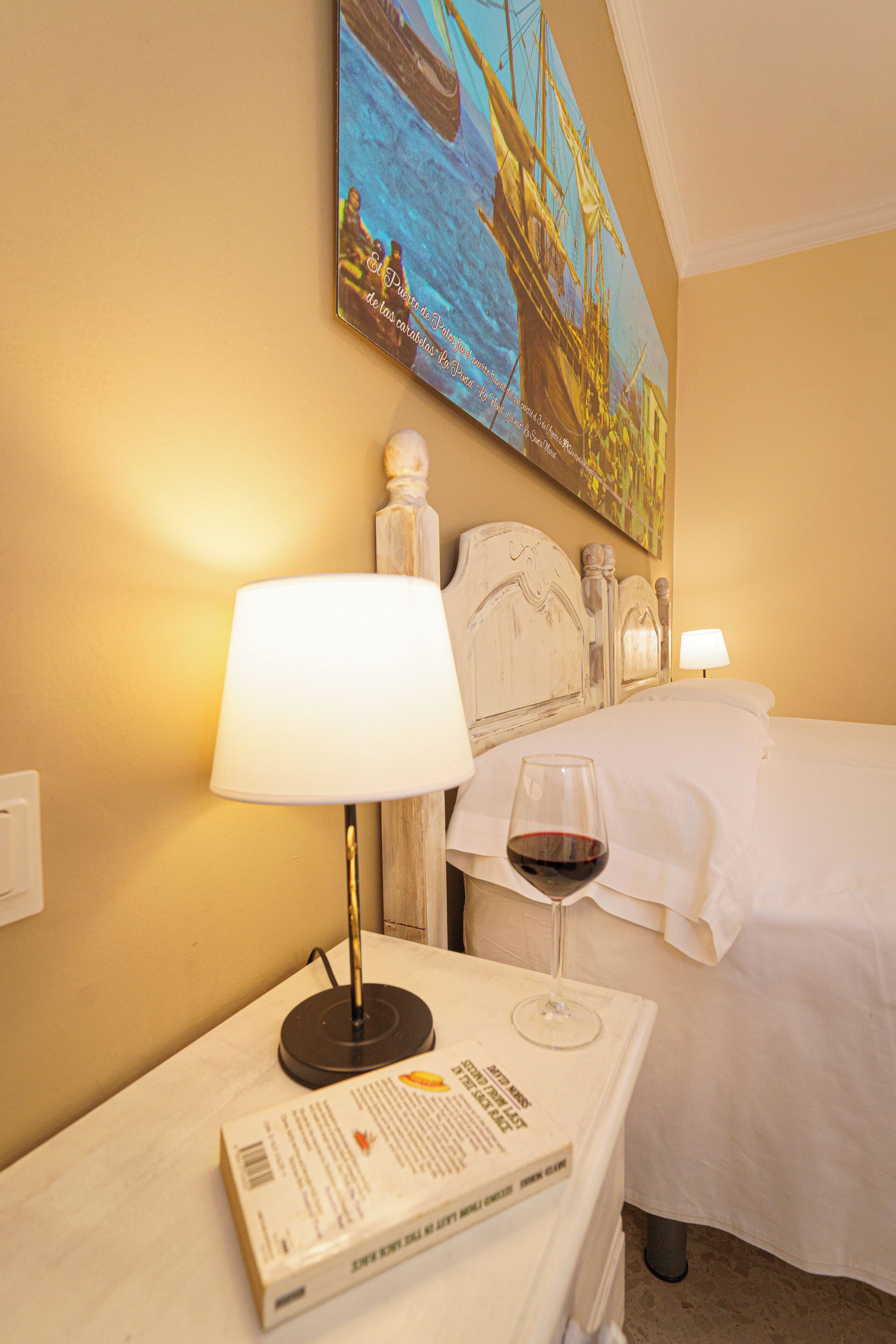 Ein Hotelzimmer mit einem Bett, einem Nachttisch mit Lampe, einem Glas Rotwein und einem Bild an der Wand.