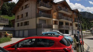A multi-story wooden building with balconies, parked cars, and a mountainous backdrop.