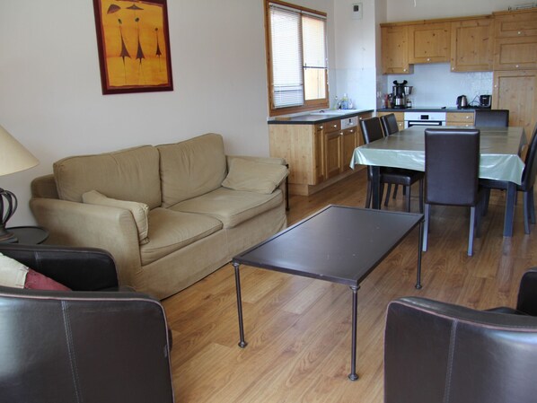 A living room with a sofa, a dining table with chairs, wooden flooring, and a kitchen area with cabinets and appliances. - Village Center Apartment with Balcony and Parking in St Jean de Sixt, Near La Clusaz (Saint-Jean-de-Sixt)
