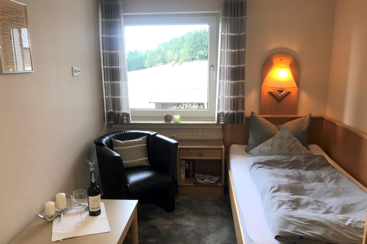 A small, cozy room with a bed, a desk with a chair, a window with curtains, and a wall-mounted lamp.