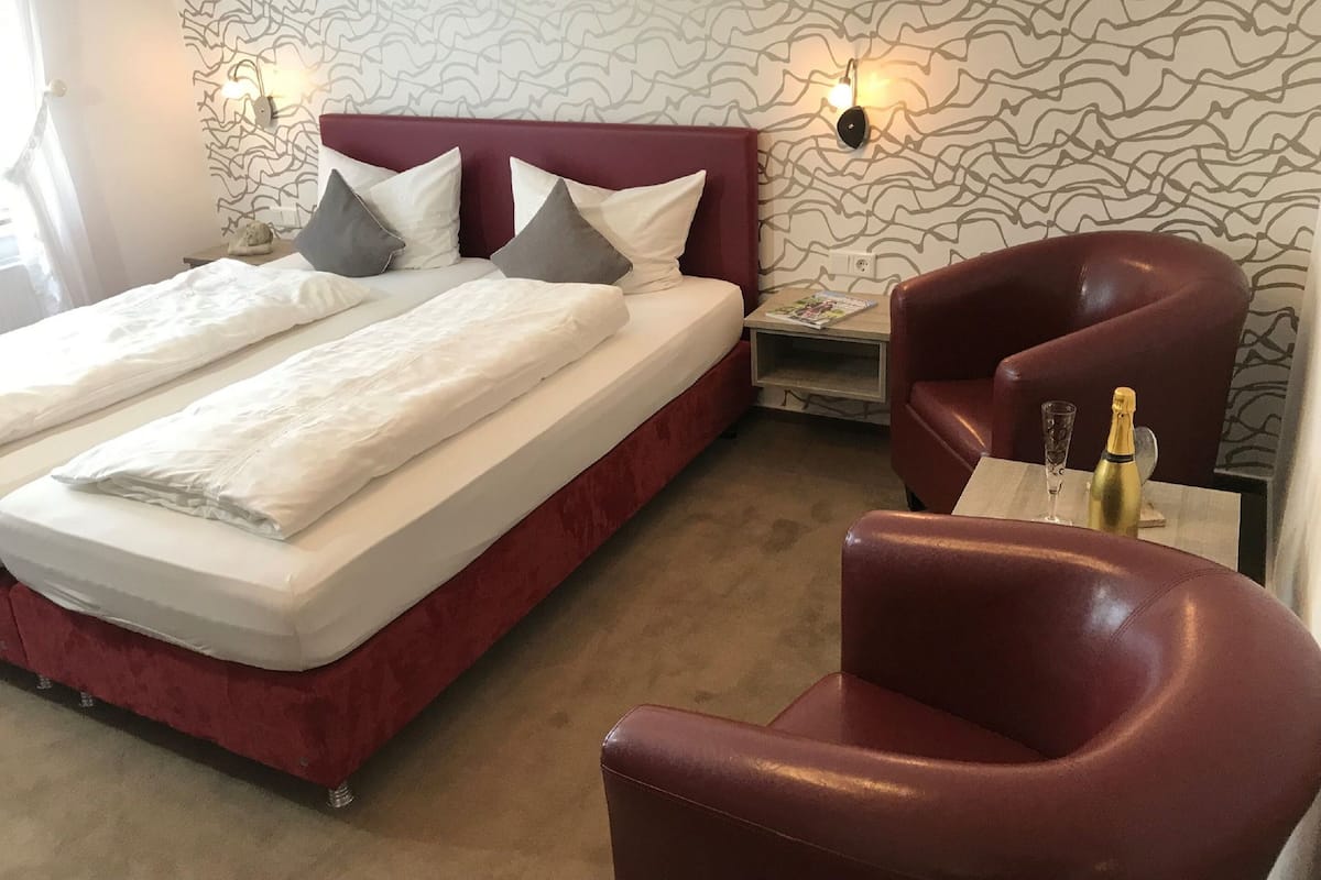 A hotel room with a bed, two pillows, a nightstand, a chair, and a bottle of champagne.