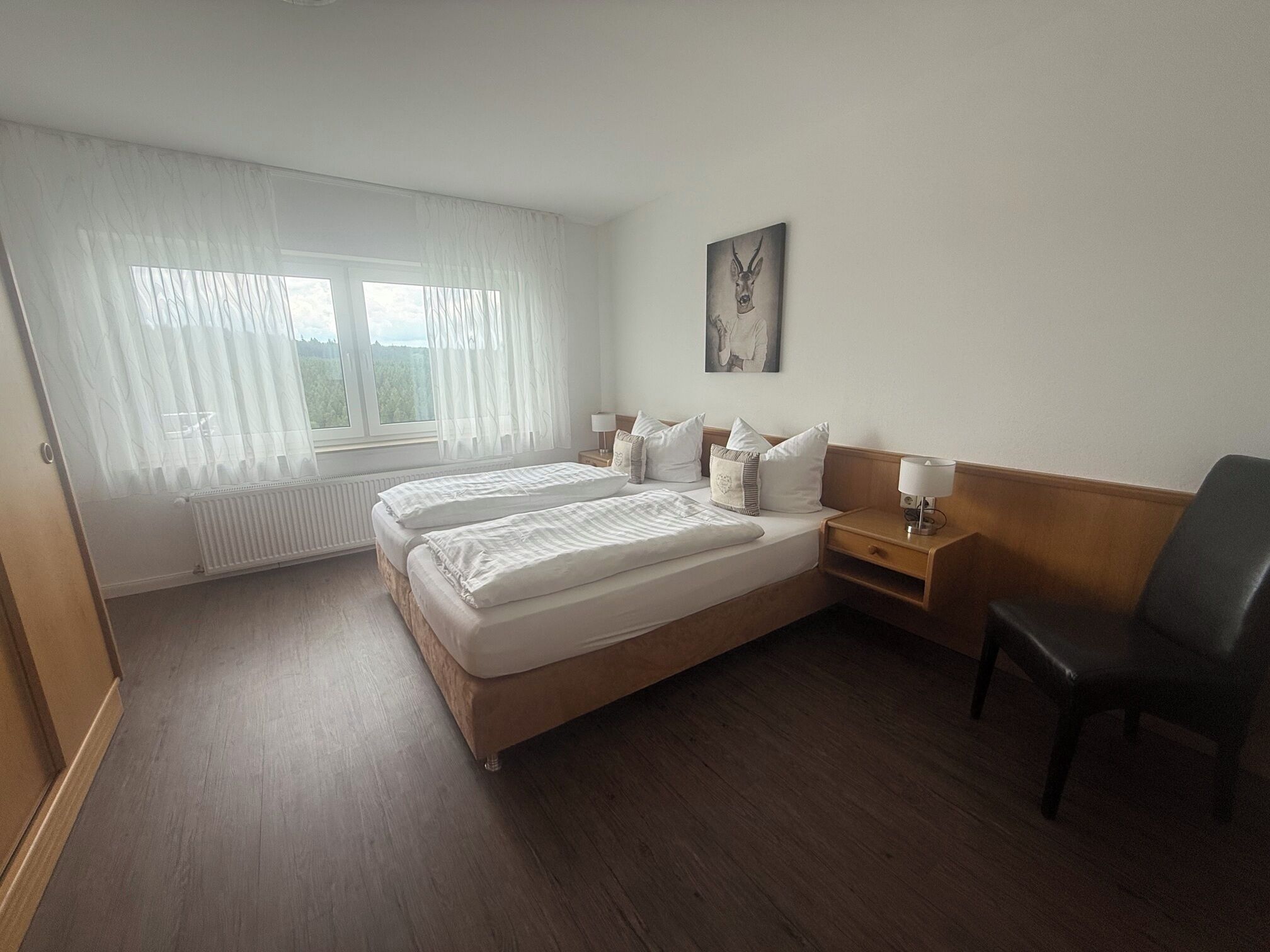 Habitación de hotel con una cama doble, una mesita de noche con lámpara, una silla, una ventana con cortinas translúcidas y un cuadro colgado en la pared.